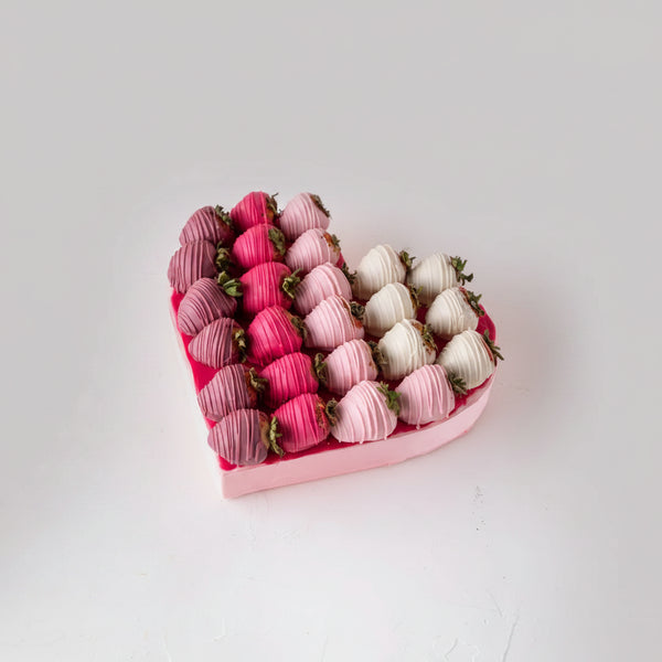 Heart-shaped box of chocolate-covered strawberries in pink and white on a light wooden surface.